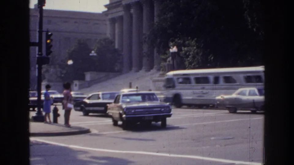 8mm home movie of a Washington DC intersection in 1975 showing a grand columned building, white bus, 1970s-era vehicles, a traffic light, and pedestrians at the crosswalk.