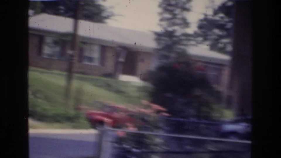 Motion-blurred 8mm frame of a brick building with green lawn, street trees, and a red car in the foreground; Washington DC area, circa 1975.