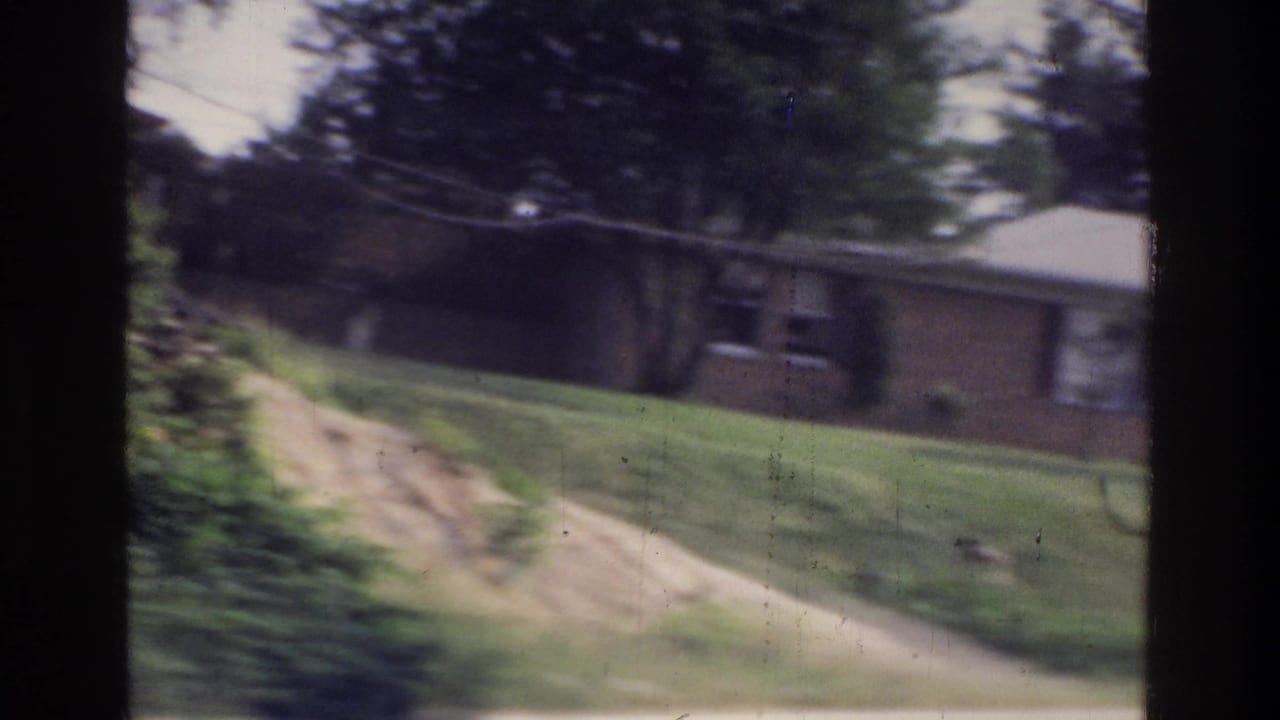 8mm home movie frame circa 1975 showing a brick ranch-style house on a sloped lawn, shot through a car window in a suburban US setting.