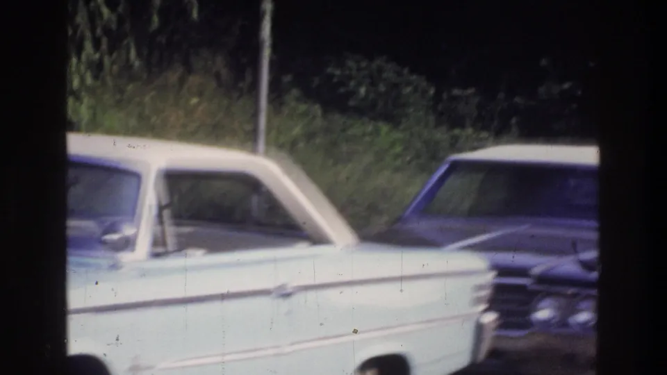 8mm film frame circa 1975 showing a pale blue large-body American sedan passing on a suburban road, with dense foliage and a utility pole visible behind it.