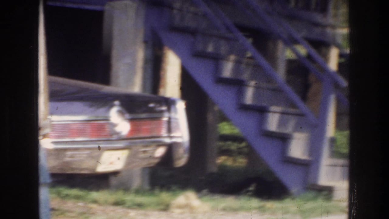 Rear view of a car parked beside a blue-painted exterior metal staircase, with green grass and a building wall visible.