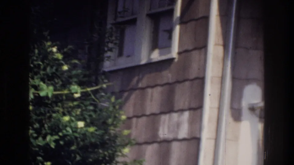 Gray-brown shingle siding on a residential building with a metal downspout and green foliage, Washington DC area, circa 1975, on 8mm home movie film.