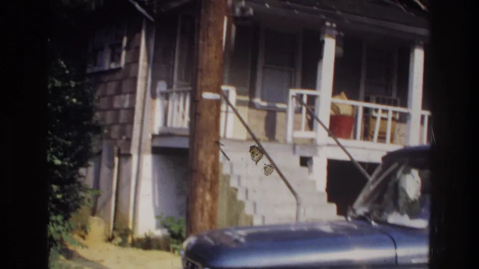 Elevated front porch with white wood railings, exterior stairs, utility pole, and blue truck partially visible at lower right, Washington DC area, circa 1975, 8mm film.
