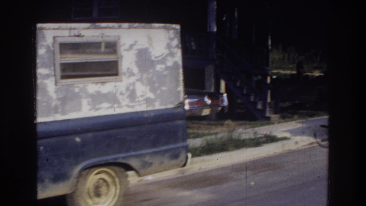 Rear of dark blue pickup truck showing peeling white utility box; a second period vehicle and sidewalk visible in background.
