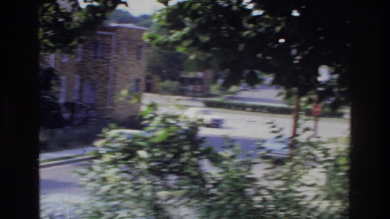 Quiet residential street with brick buildings and mature trees, shot from elevated angle, 8mm footage.