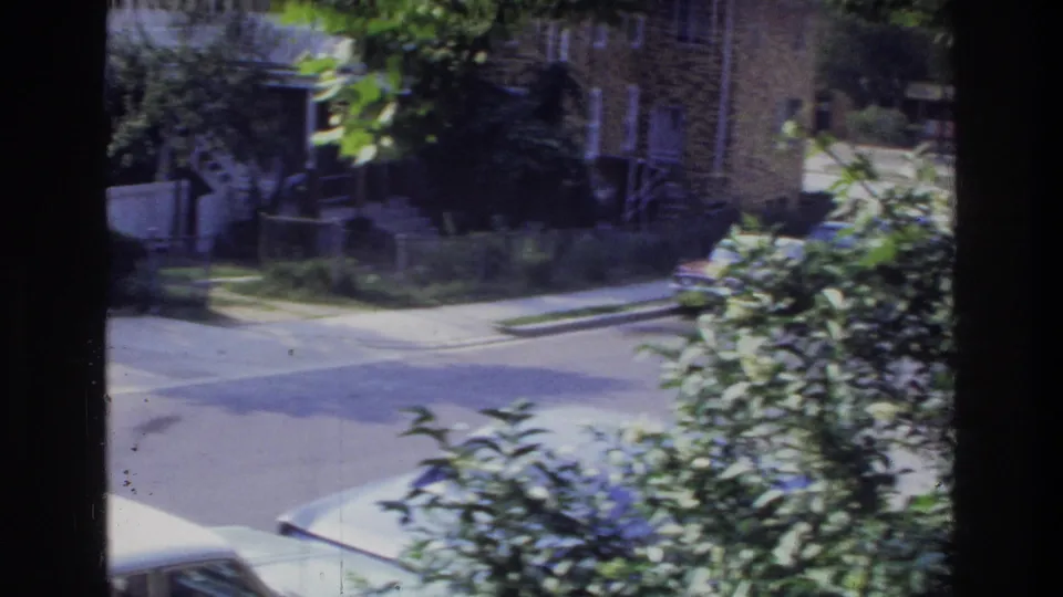 Brick suburban house with garden vegetation and sidewalk in foreground, 1970s American residential neighborhood, 8mm home movie.