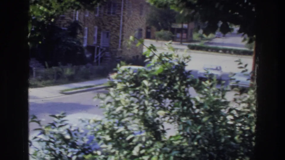 Suburban brick house and residential street viewed past garden foliage, 1970s American neighborhood, 8mm home movie.
