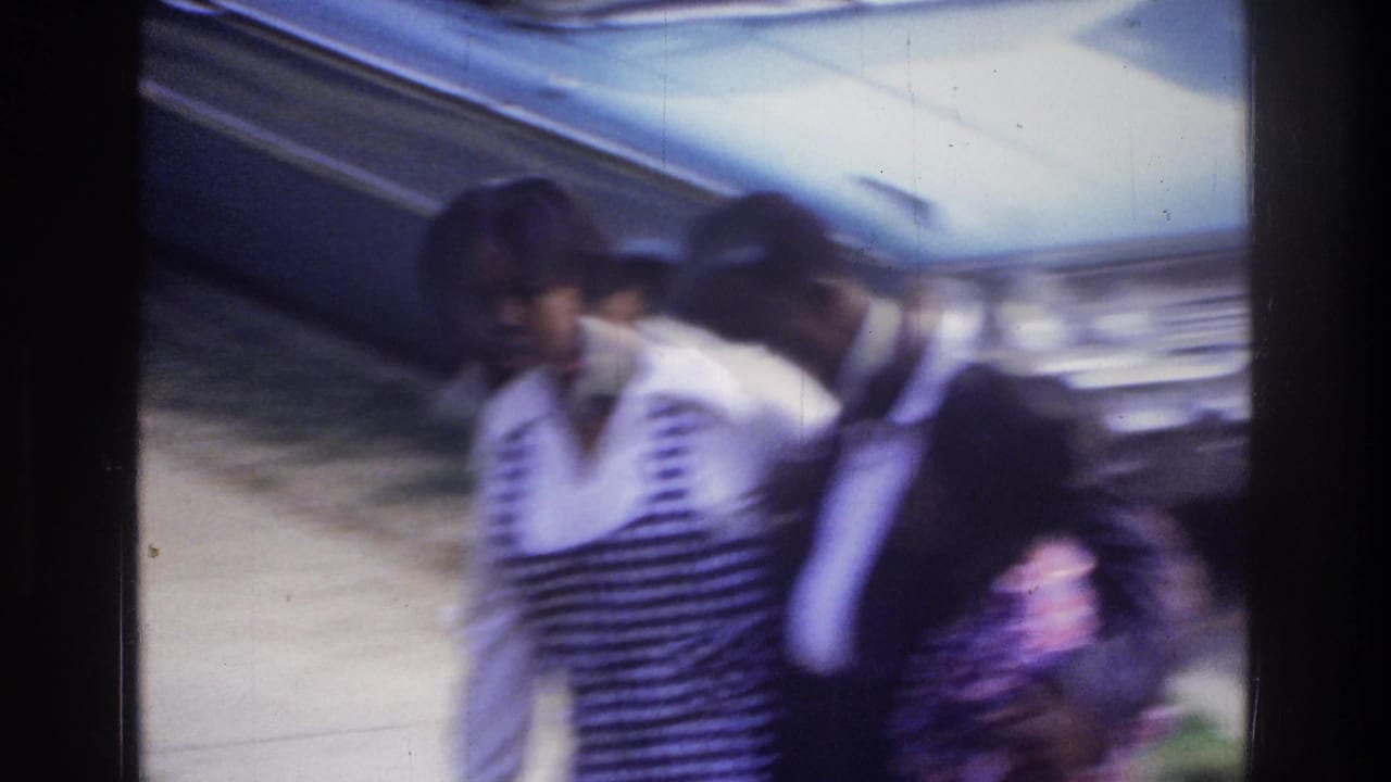 Heavy motion blur in 8mm home movie frame showing a family group — woman in striped top, man in dark jacket, child — near a 1970s-era American car.