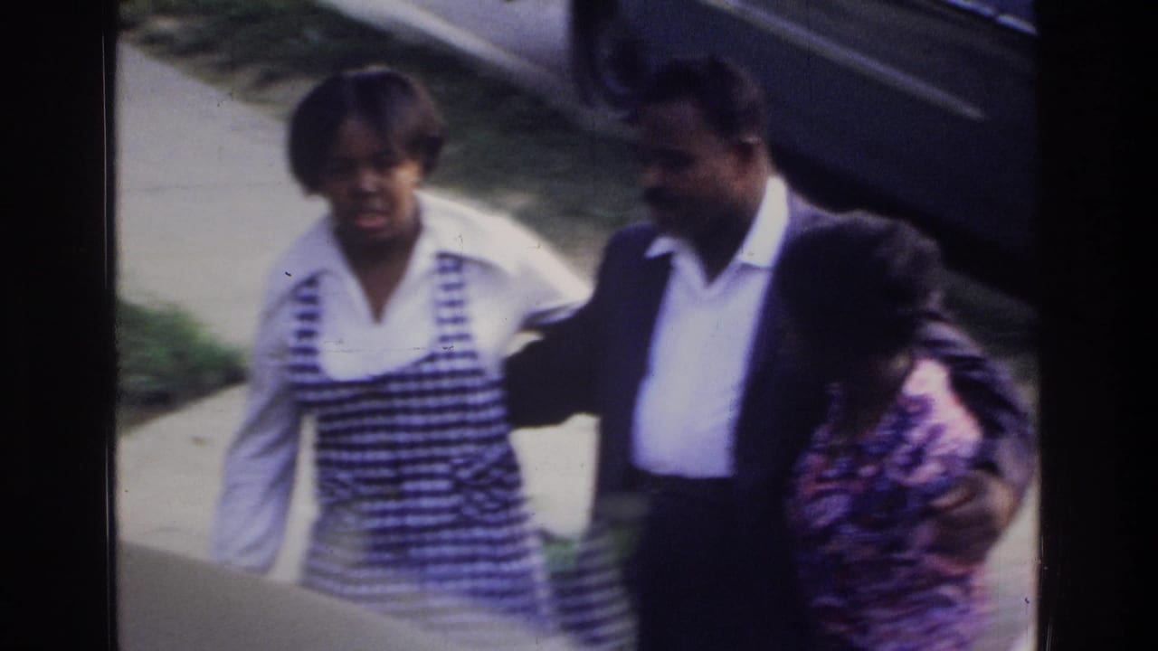 Woman in striped top and man in dark blazer with a child on a concrete sidewalk beside green shrubbery.