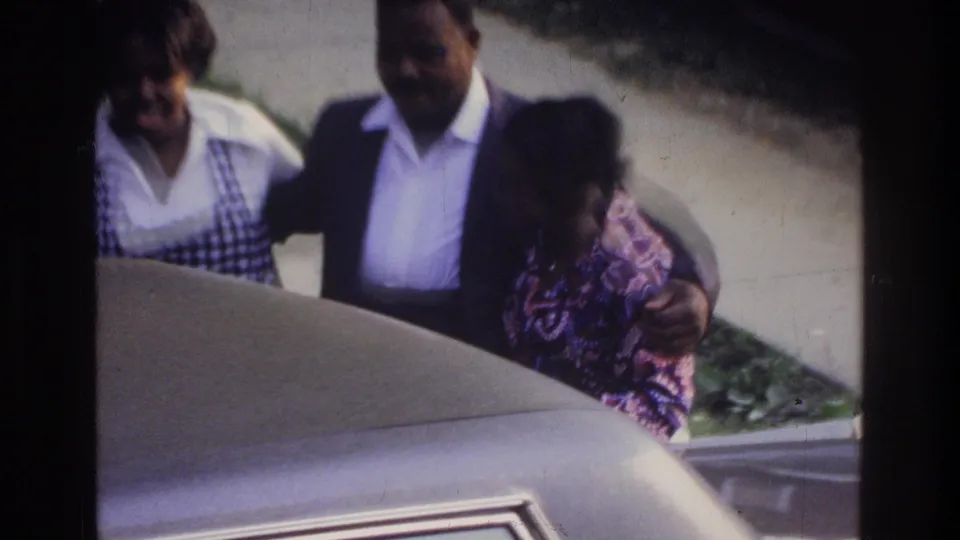 Woman in striped top and man in dark jacket with a child, positioned beside the hood of a large light-colored 1970s American sedan; green foliage behind them.