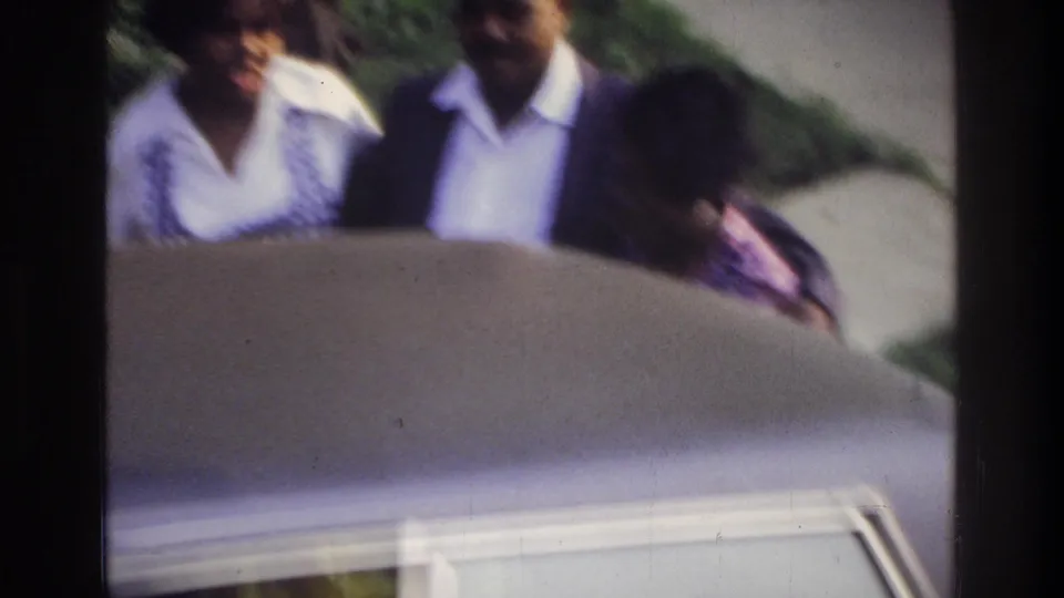 Motion-blurred 8mm home movie frame showing two adults and a child near the hood of a large light-colored American car, green foliage in background.