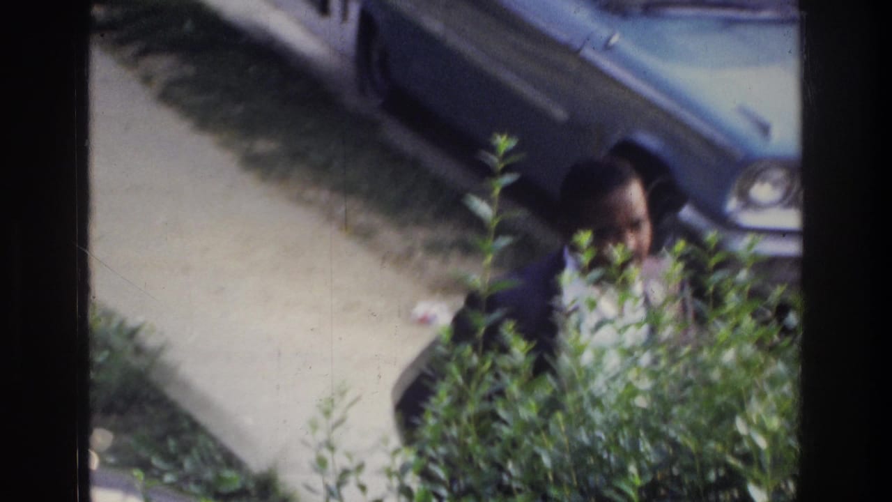 Well-dressed Black man in dark suit approaches a large American sedan on a driveway, framed through foliage; Washington DC home movie, circa 1975.