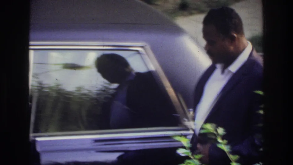 Well-dressed Black man opens the door of a large dark American sedan, framed through foreground bushes; Washington DC home movie, circa 1975.