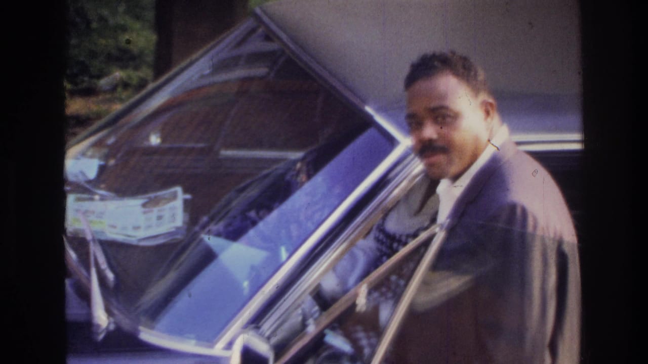 In 1975 Washington, DC home-movie footage, a suited man stands by the open driver door of a classic sedan.