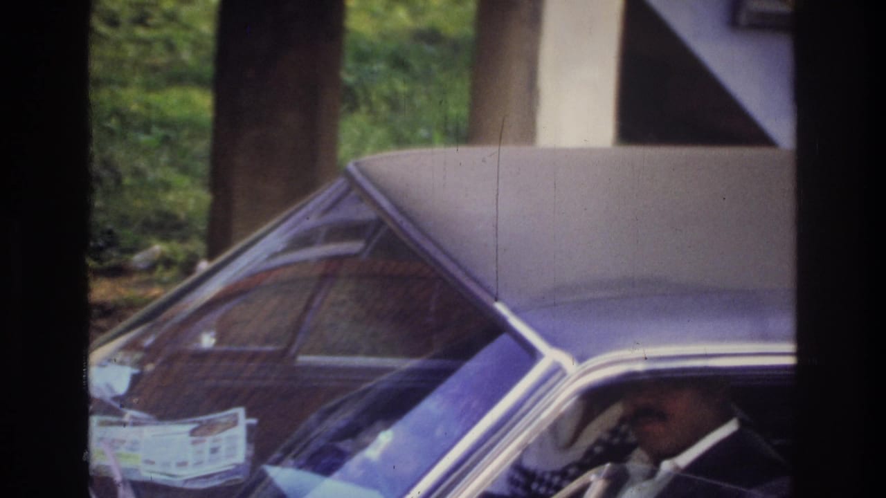 Man sits behind the wheel of a classic sedan with paper on the dashboard.
