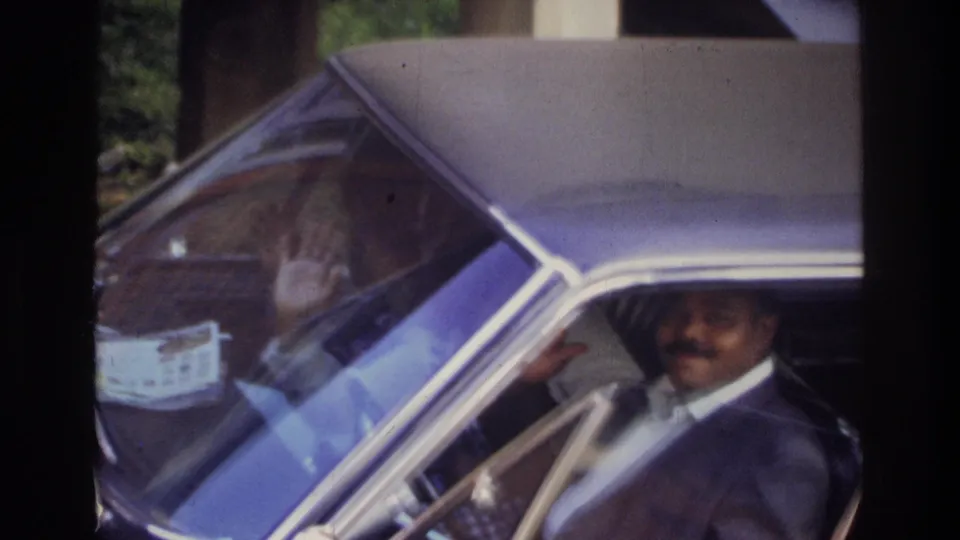 The suited man smiles and waves from the driver's seat of the sedan in 1975 Washington, DC home-movie footage.
