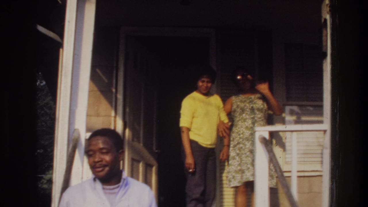 A man in a light shirt stands in the foreground while two adults stand on the porch behind him.