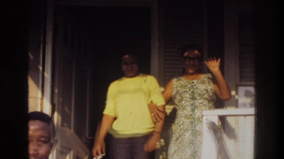 1975 Washington, DC 8mm frame showing a woman waving beside another adult on a residential porch.