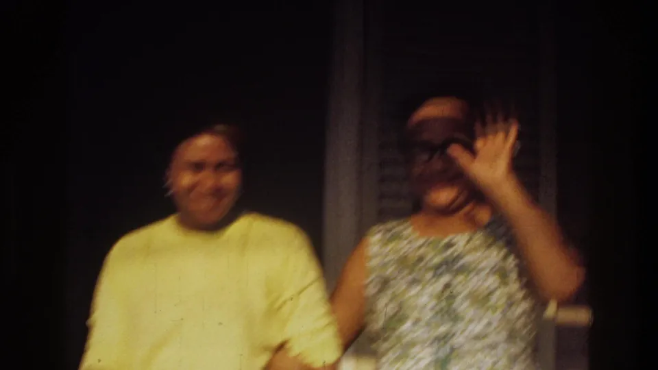 Soft-focus 1975 Washington, DC home-movie frame with two adults on a porch and a woman waving near the railing.