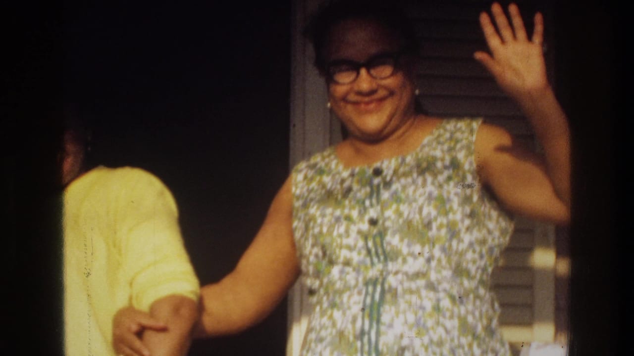 1975 Washington, DC home-movie frame of a smiling woman in glasses waving near a doorway while another person in yellow is partly visible.