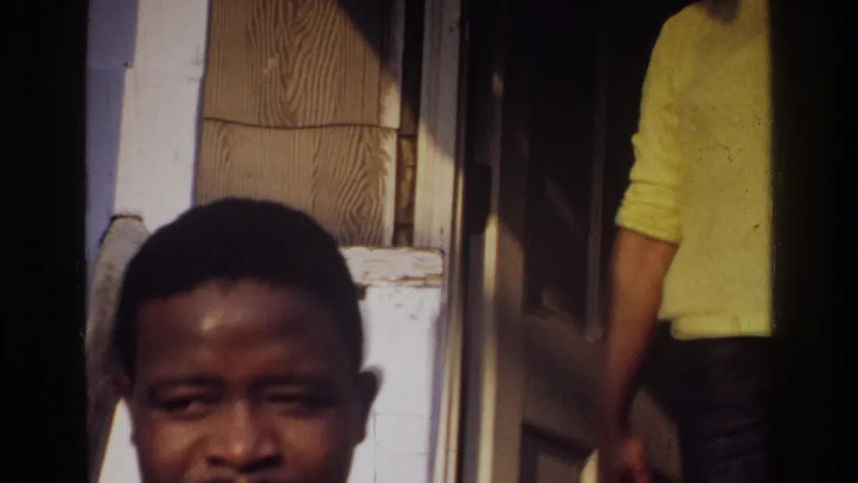 1975 Washington, DC home-movie frame with a young man close to the camera near a house entrance and a partly visible person in yellow.