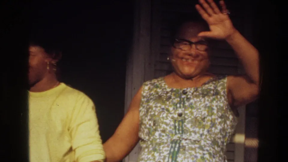 1975 Washington, DC home-movie frame of two women at a doorway, with one turning aside and another smiling while raising her hand.