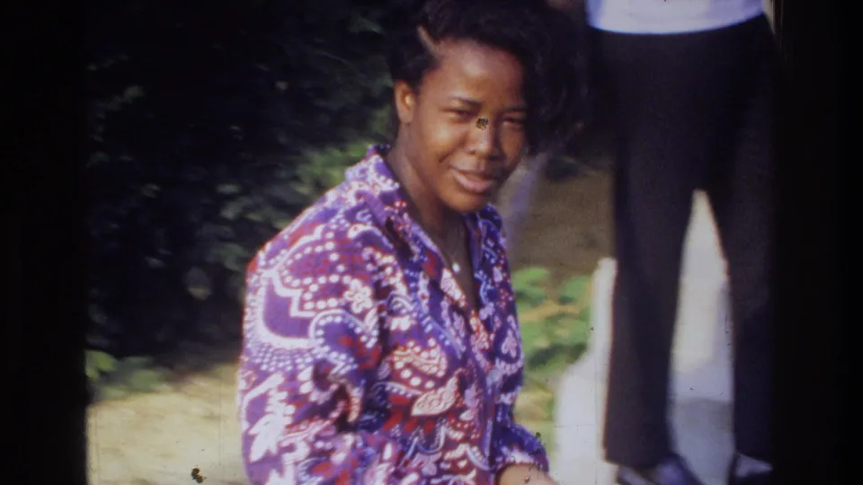 1975 Washington, DC home-movie frame of a young man in a purple paisley shirt looking toward the camera with greenery and a partial standing figure behind him.
