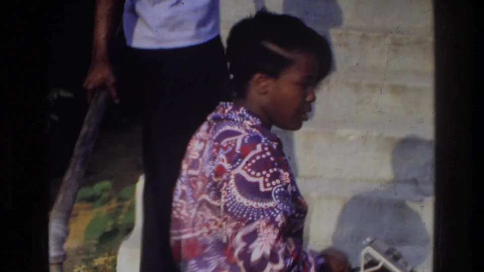 In 1975 Washington, DC home-movie footage, a woman in a patterned blouse moves past porch steps with another person partly visible nearby.
