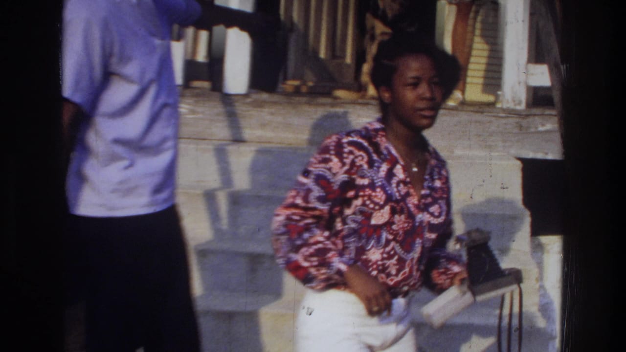 Woman in patterned blouse and white pants holds a book near a porch.