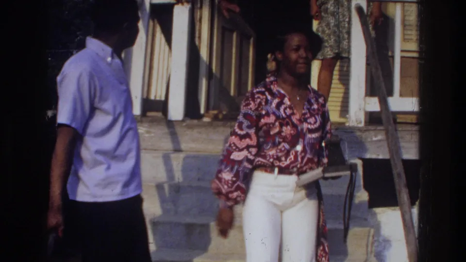 A woman in a patterned blouse approaches the porch as a man in a light shirt stands partly in the foreground in the 1975 Washington, DC scene.