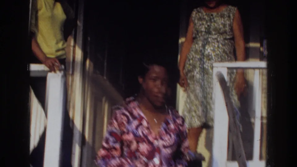 The woman moves near the porch railing as other people stand around the doorway, including a woman in a patterned dress.