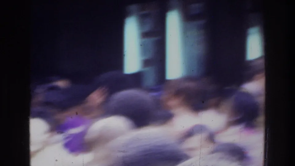 Blurred 1975 home-movie view of a crowded gathering, with people moving through a dim or shaded space.