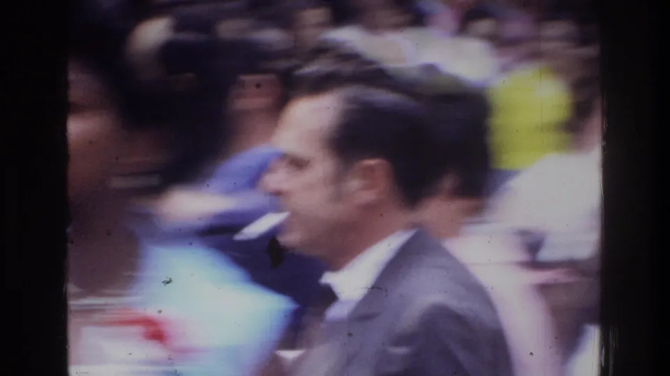 1975 Washington, DC home-movie frame showing a suited man in profile amid a blurred crowd of people.