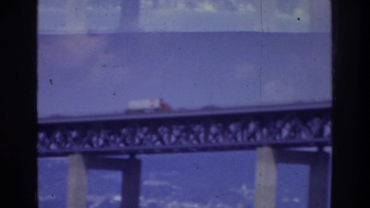 1975 Washington, DC home-movie frame showing a white truck on a steel truss bridge above water, with dark window edges in the foreground.