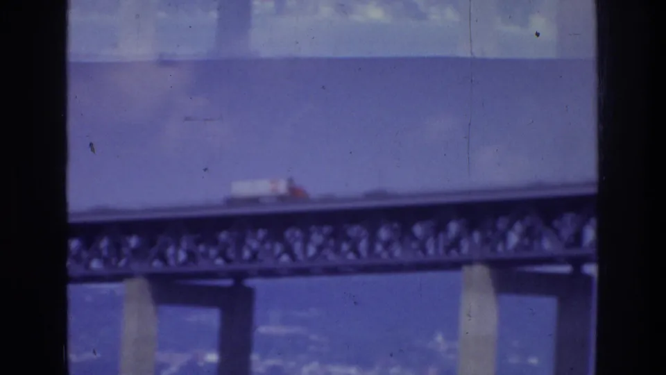 1975 Washington, DC home-movie frame showing a white truck on a steel truss bridge above water, with dark window edges in the foreground.