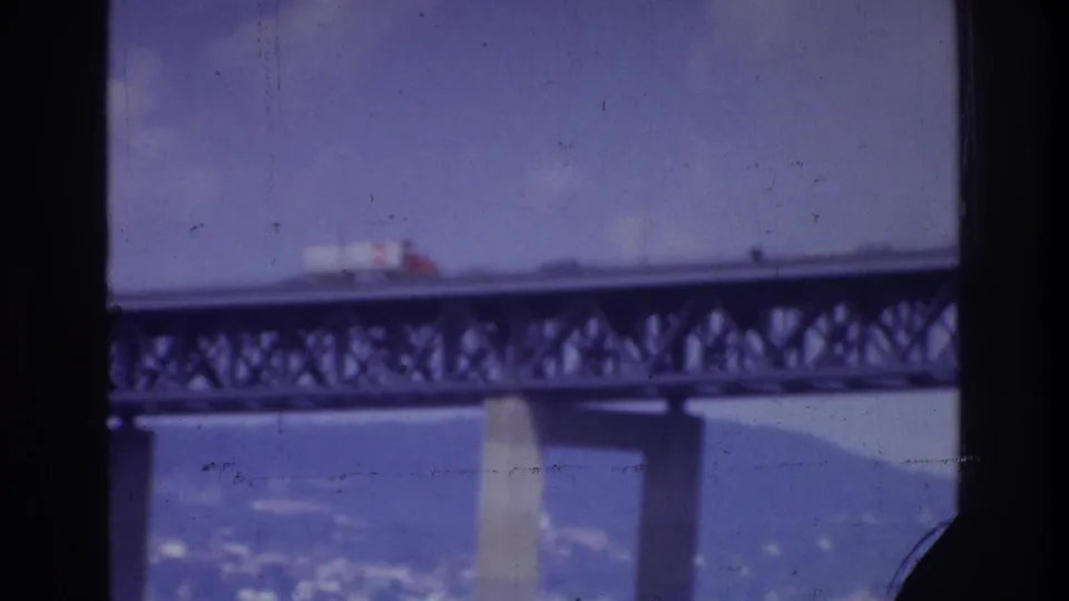 A white truck crosses the bridge span in the supplied 1975 Washington, DC footage, partly obscured by a dark vertical window frame.