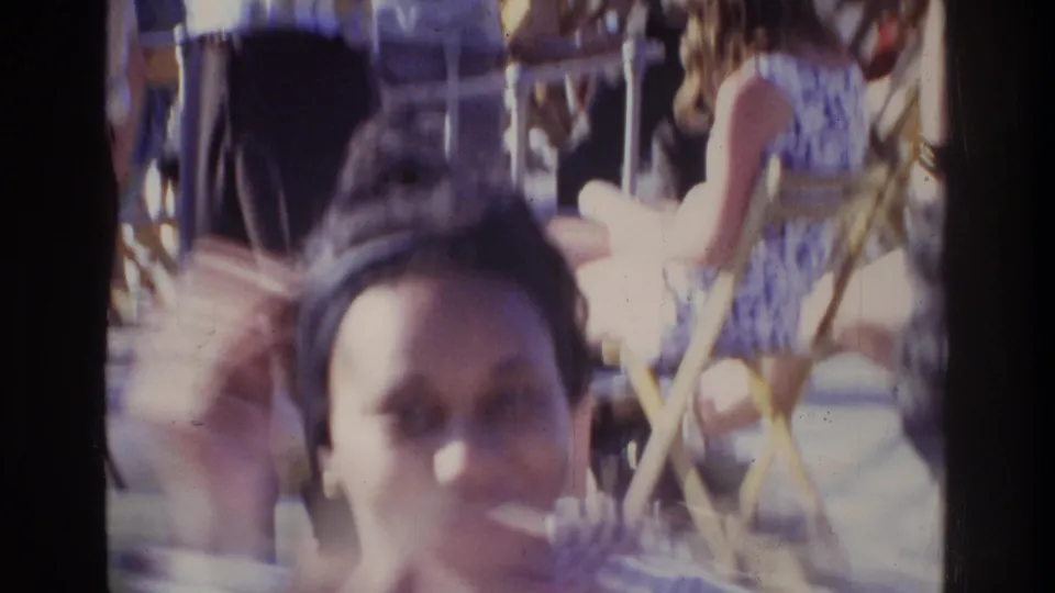 Woman smiling toward camera at an outdoor 1975 gathering; gold-framed folding chairs and other attendees visible in the background.