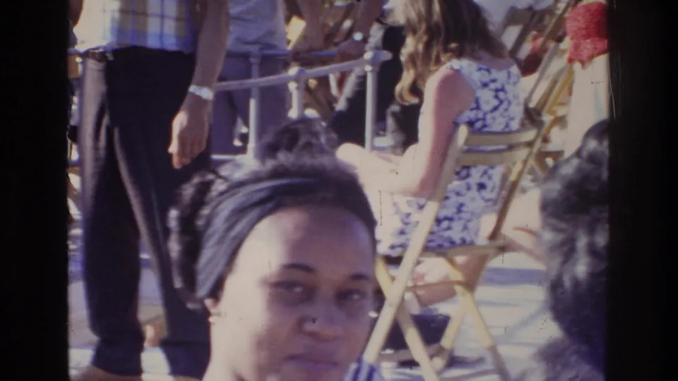 Woman wearing a headband at an outdoor 1975 gathering in Washington DC; a man in dark trousers and a seated woman in a floral top visible with gold-framed folding chairs behind.