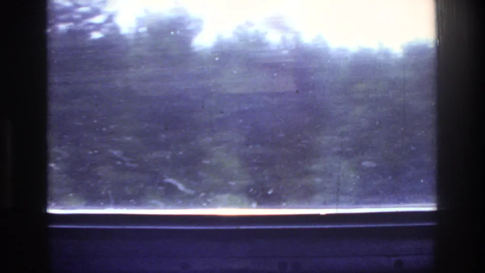 1975 8mm road footage: blurred roadside foliage and bright sky through a moving vehicle window, with orange-red light glare at the sill and purple-blue film cast.