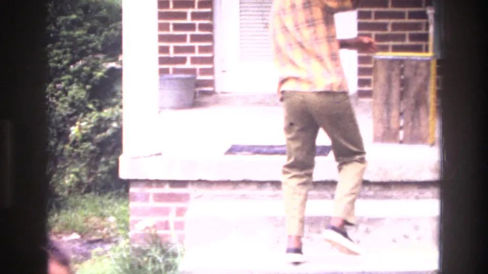 1970s 8mm home movie frame: a young person in a plaid shirt and olive trousers seen from behind, standing on concrete steps beside a brick row-house facade with green foliage.