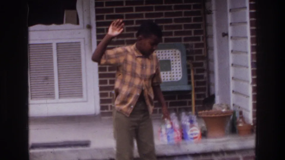 1970s 8mm home movie: a young person in a plaid shirt gestures with a raised hand outside a brick row house, with a terracotta flowerpot, bottles, and a louvered vent panel visible.