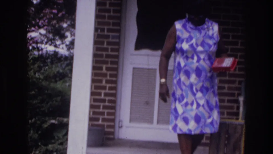 Woman in mid-1970s geometric sleeveless shift dress holding a small red box outside a brick suburban house, 8mm home movie, Washington DC area, c. 1975.