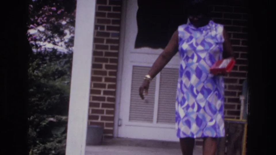 Woman in mid-1970s geometric-print dress walking with red box outside suburban brick home, 8mm home movie, Washington DC area, c. 1975.