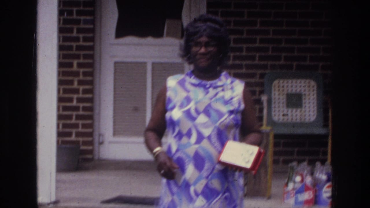 Mid-1970s 8mm home movie still: a woman in a geometric-print sleeveless dress stands on a brick porch holding a red-bordered item, smiling, Washington DC area, circa 1975.