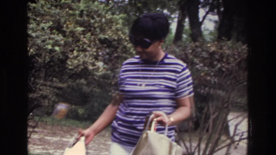 Mid-1970s 8mm home movie still: a woman in a striped shirt and sunglasses walks through a residential garden carrying a beige tote bag, Washington DC area, circa 1975.