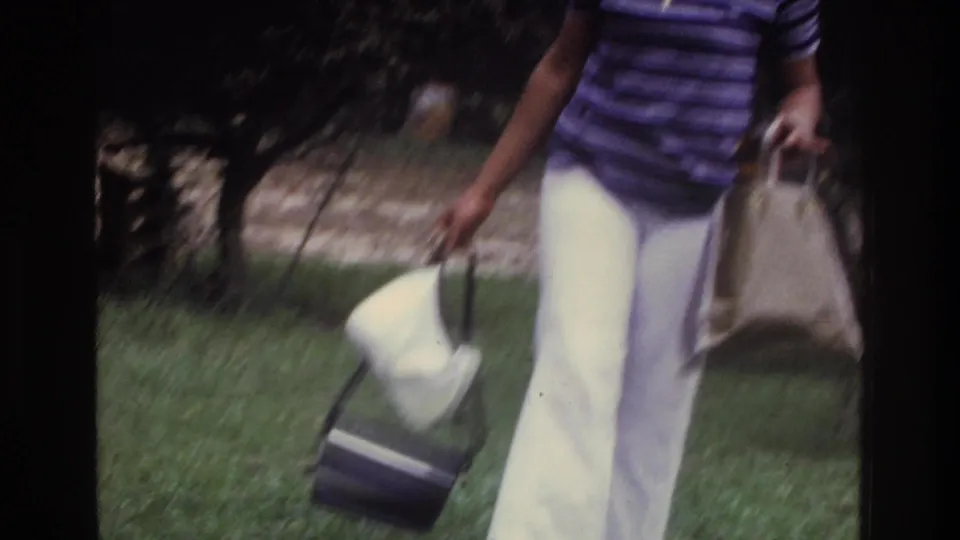 Mid-1970s 8mm home movie still: a woman in wide-leg white trousers and a striped shirt walks across a residential lawn carrying a tote bag and white hat, Washington DC area, circa 1975.