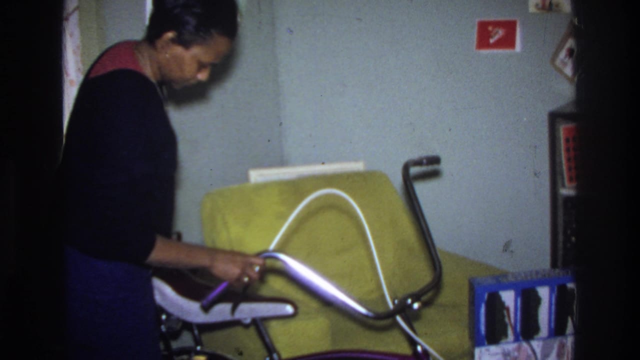 Woman in dark sweater with red collar adjusting chrome ape-hanger handlebars of a purple bicycle inside a US home interior, circa 1975