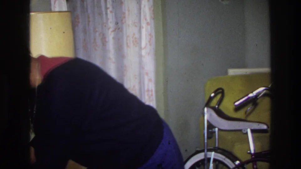 Purple banana-seat bicycle with white seat and chrome ape-hanger handlebars in a living room; woman partially visible bending behind it, patterned curtains and yellow lamp at left, circa 1975