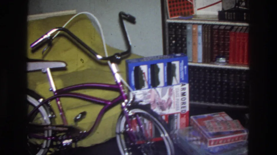 Purple banana-seat bicycle with chrome handlebars and white seat in a 1970s living room; bookshelf at right holds stacked boxes including one with legible text ARMORED, circa 1975