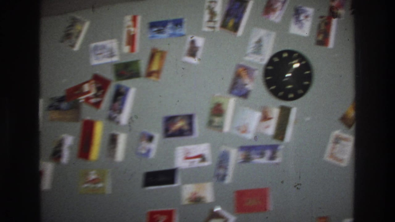 A light-green interior wall covered with colorful Christmas cards and a round black analog clock, from a 1975 Washington DC home movie.
